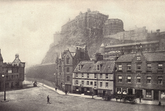 Black and white photograph of Edinburgh Castle in Scotland, on a hill surrounded with haze, taken from below that includes a clear view of a street and buildings, taken in the late 19th century.